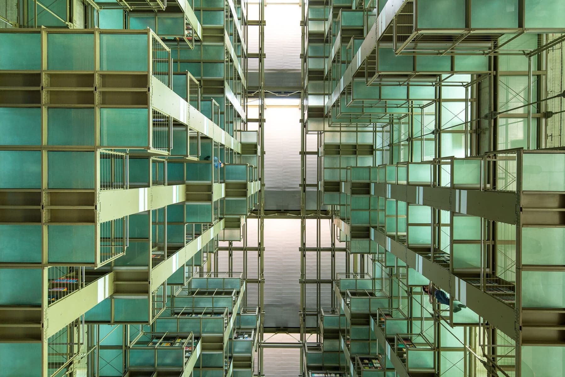 Looking straight up through tiers of green glass and steel book stacks toward the skylight